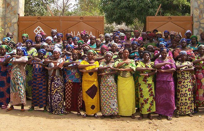 Group of women in colorful dresses stand in front of brown gate, arms crossed in solidarity, outdoors with a sign reading 