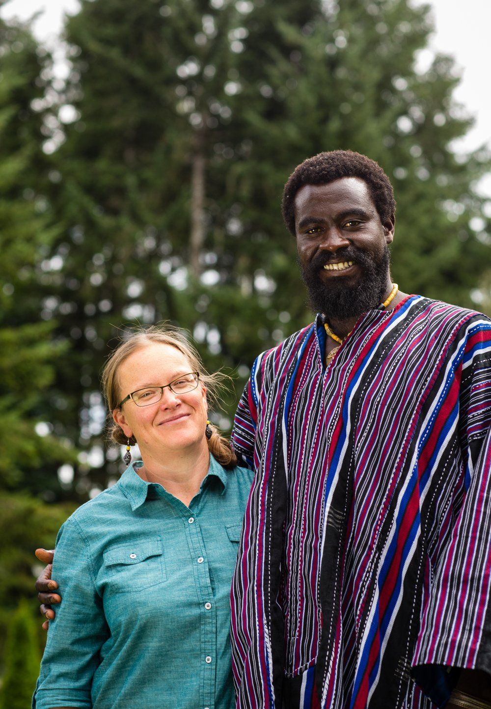A smiling Black man in colorful robes and a white woman with glasses pose together outside, trees in the background.