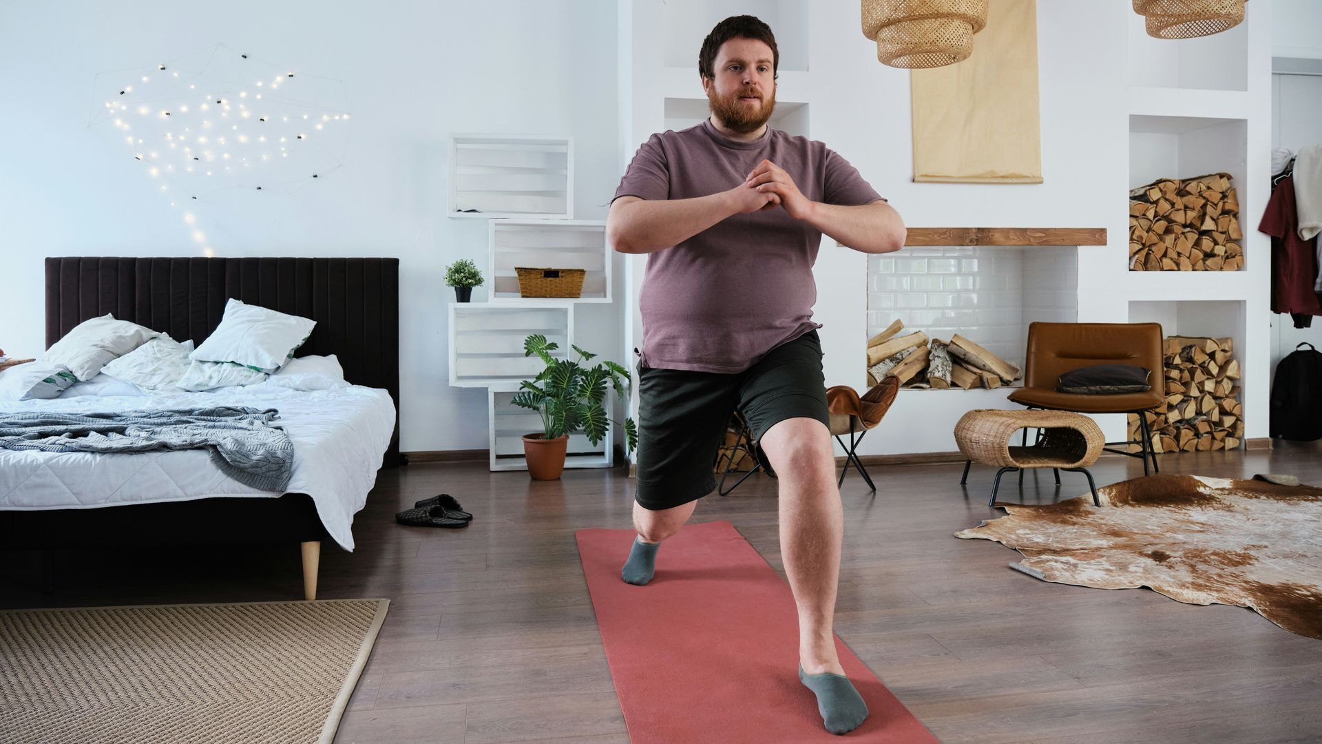 A man in a home gym doing a lunge on a red mat, facing the camera. He wears shorts and a tee-shirt.