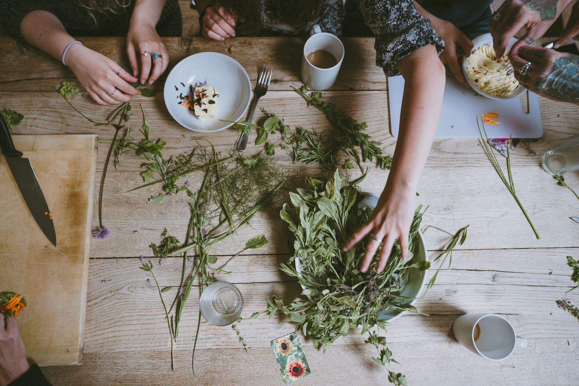 Overhead shot of a wooden table with people arranging herbs and food. Hands reach for fresh greenery and plates of food.