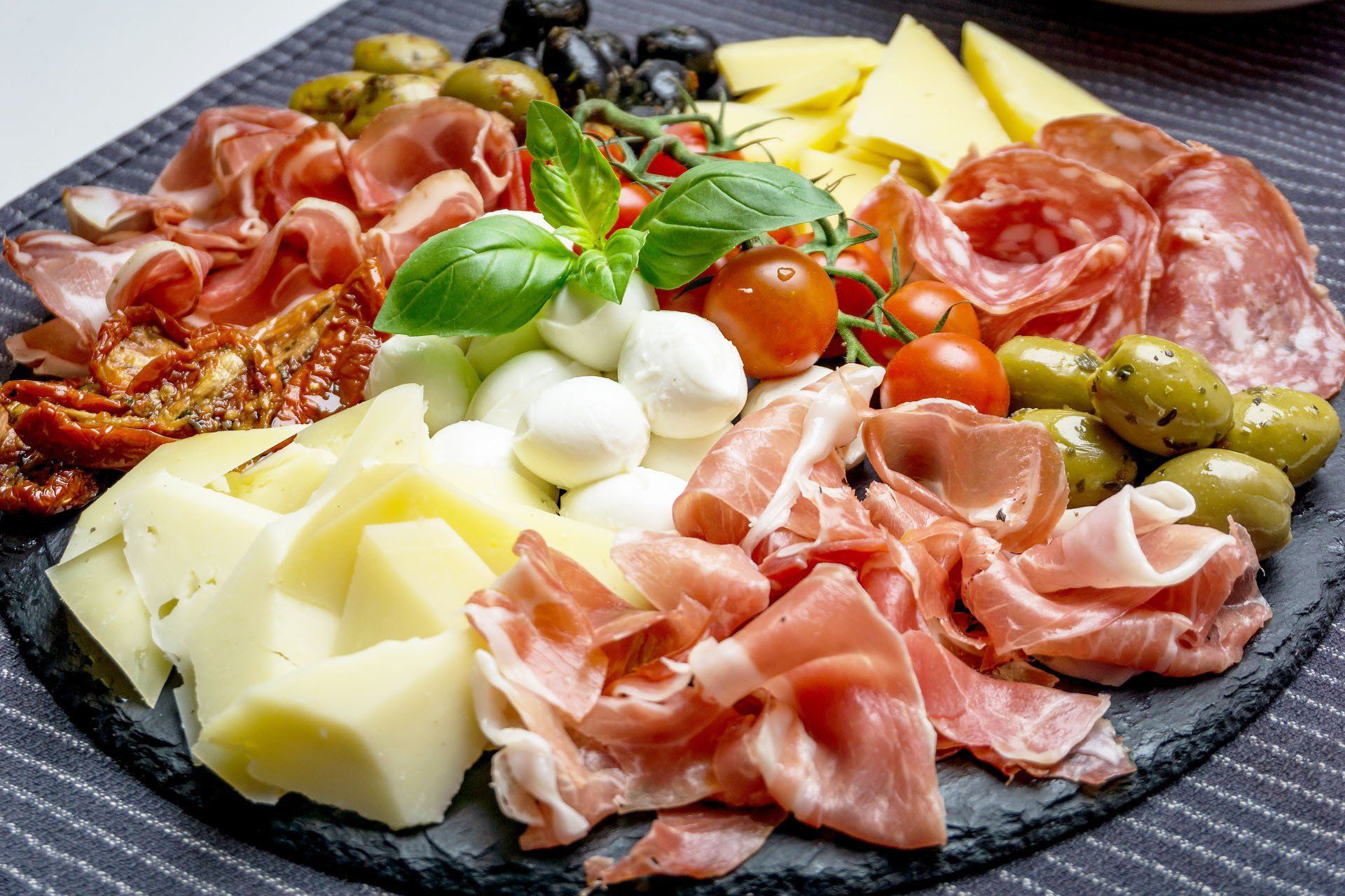 Charcuterie board with meats, cheeses, olives, mozzarella balls, tomatoes, and basil on a dark platter.