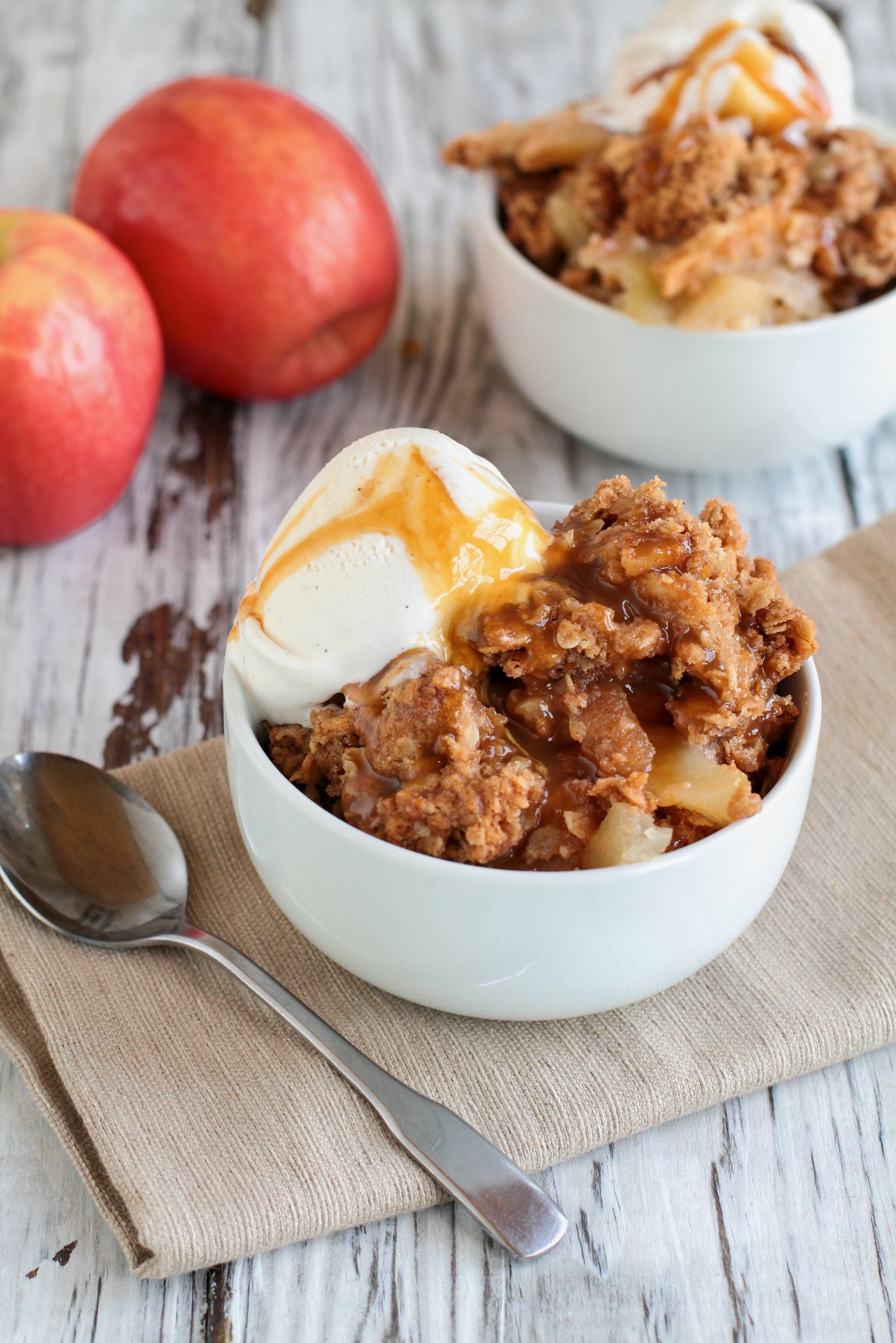 Apple crisp with ice cream and caramel sauce in a white bowl, served on a burlap napkin with a spoon. Red apples in the background.