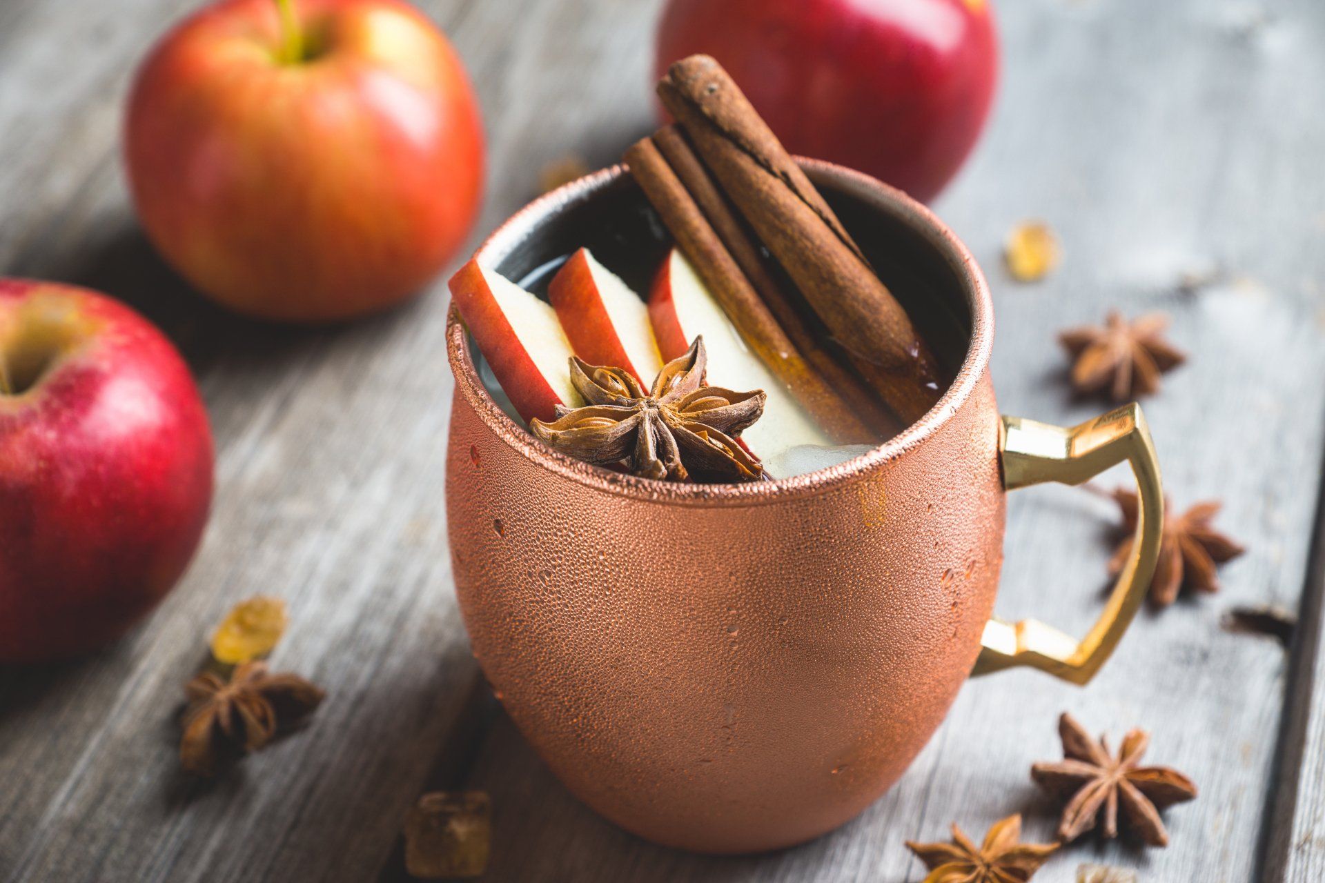 Copper mug with apple slices, cinnamon sticks, and star anise, garnished with red apples on a wooden surface.
