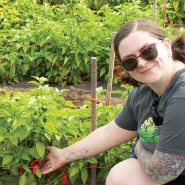Woman with sunglasses smiles, pointing at chili peppers growing in a field. She is surrounded by lush green plants.