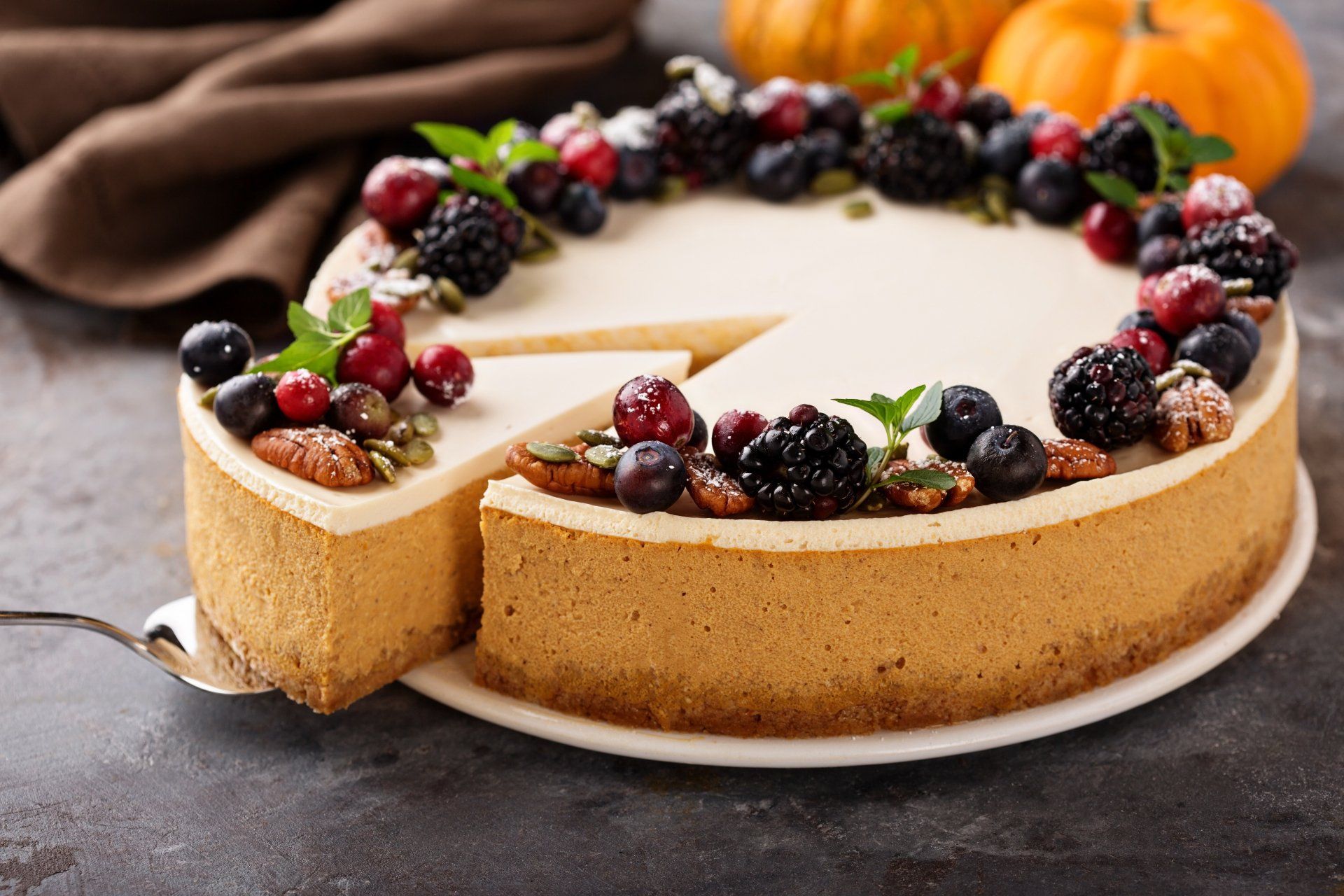 Pumpkin cheesecake topped with berries, nuts, and mint, with a slice being removed.
