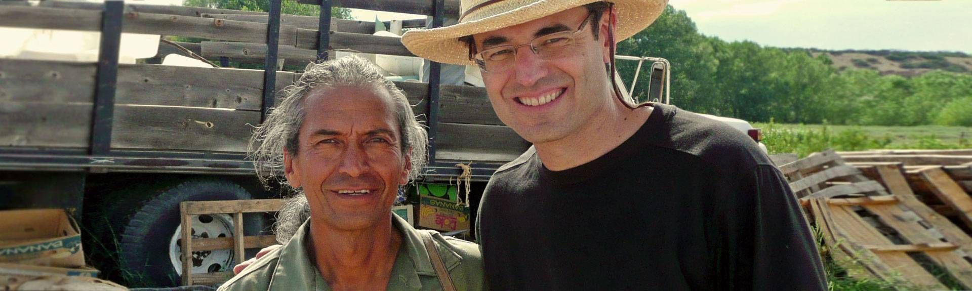 Two men pose for a photo next to a truck bed full of wooden planks. One man is older with gray hair. The other wears a straw hat and glasses.