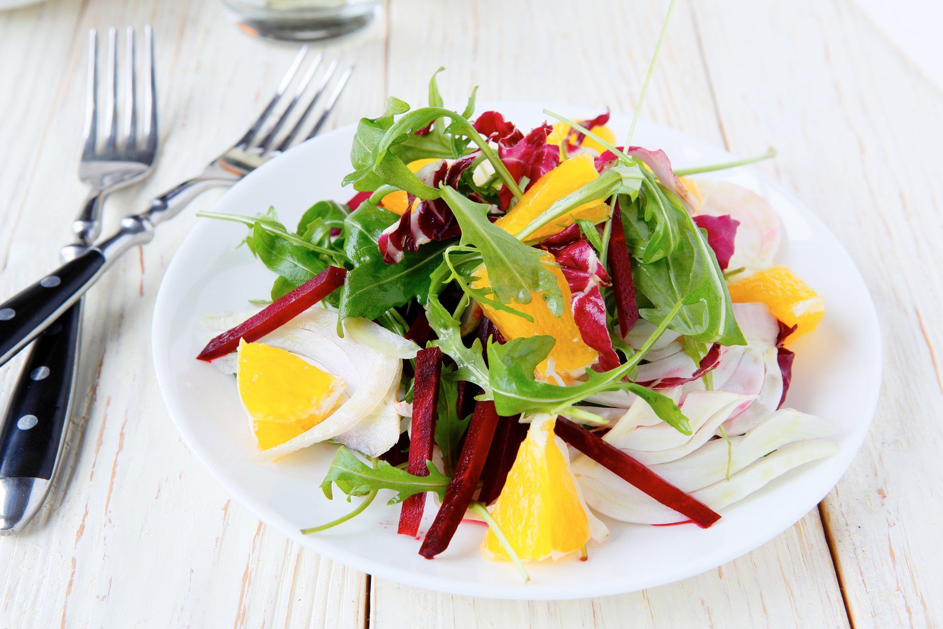 A vibrant salad on a white plate. It includes orange slices, dark red beets, and green leafy vegetables on a white wooden table.