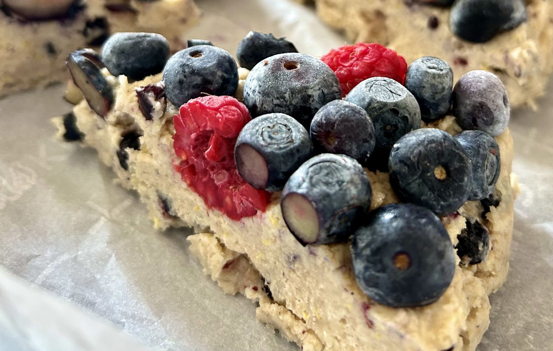 Close-up of a blueberry and raspberry scone on parchment paper. The baked good is light-colored and loaded with fresh, frosted fruit.