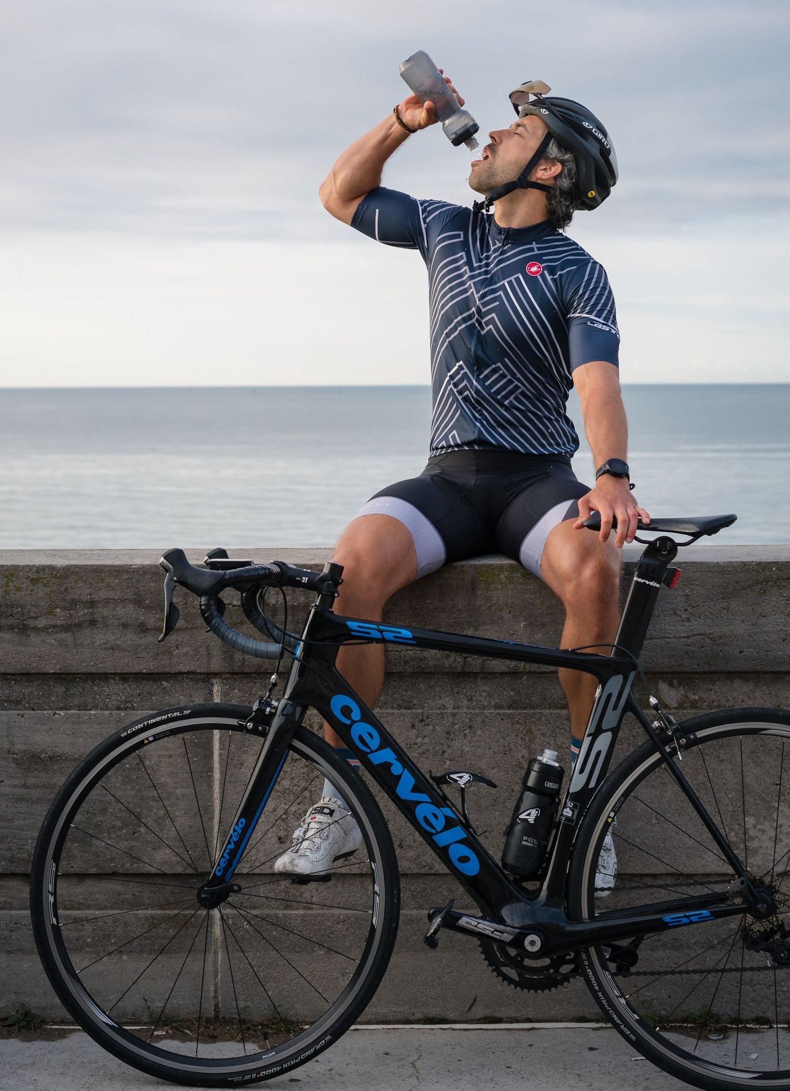 Cyclist on a seawall drinking from a bottle. He's wearing a helmet, cycling jersey and shorts. His bike is beside him with the ocean in the background.