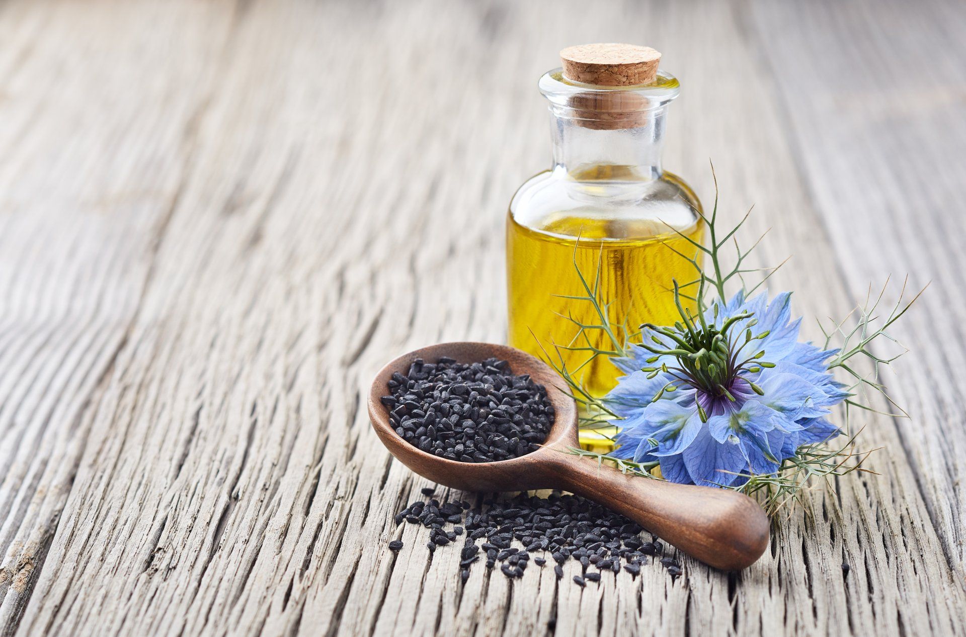 Bottle of black seed oil, wooden spoon with black seeds, blue flower on a weathered wooden surface.