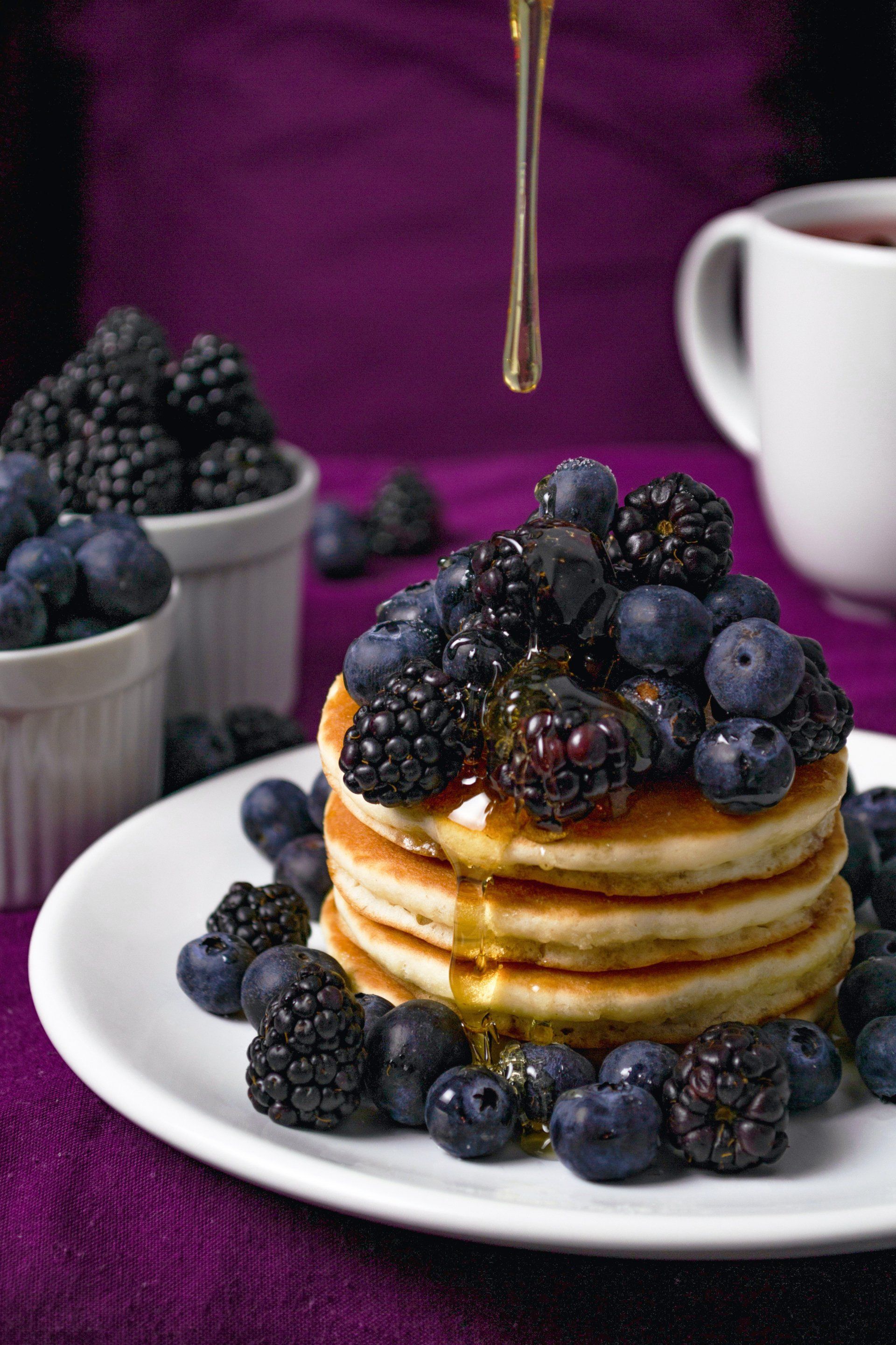 Stack of pancakes topped with blueberries, blackberries, and syrup being drizzled on a white plate, purple background.