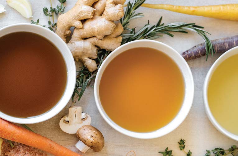 Three bowls of broth with ginger, herbs, carrots, and a mushroom scattered around.