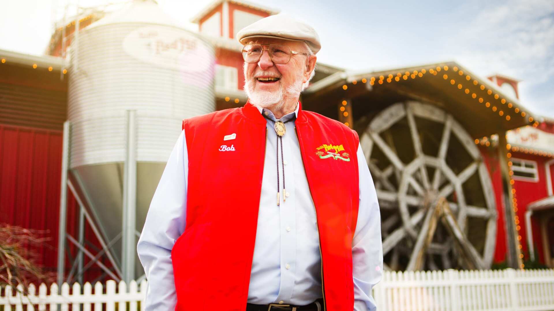Man in red vest and white hat smiles outside a red barn with a water wheel.