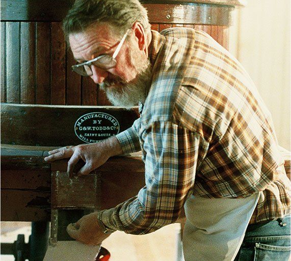 Elderly man with a white beard and glasses working on a wooden wine press in a rustic setting; he's wearing a plaid shirt and apron.