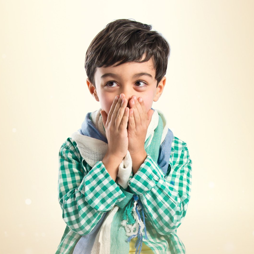 A young boy with dark hair covers his mouth with his hands, looking to the side in surprise; he wears a green plaid shirt and a patterned scarf.