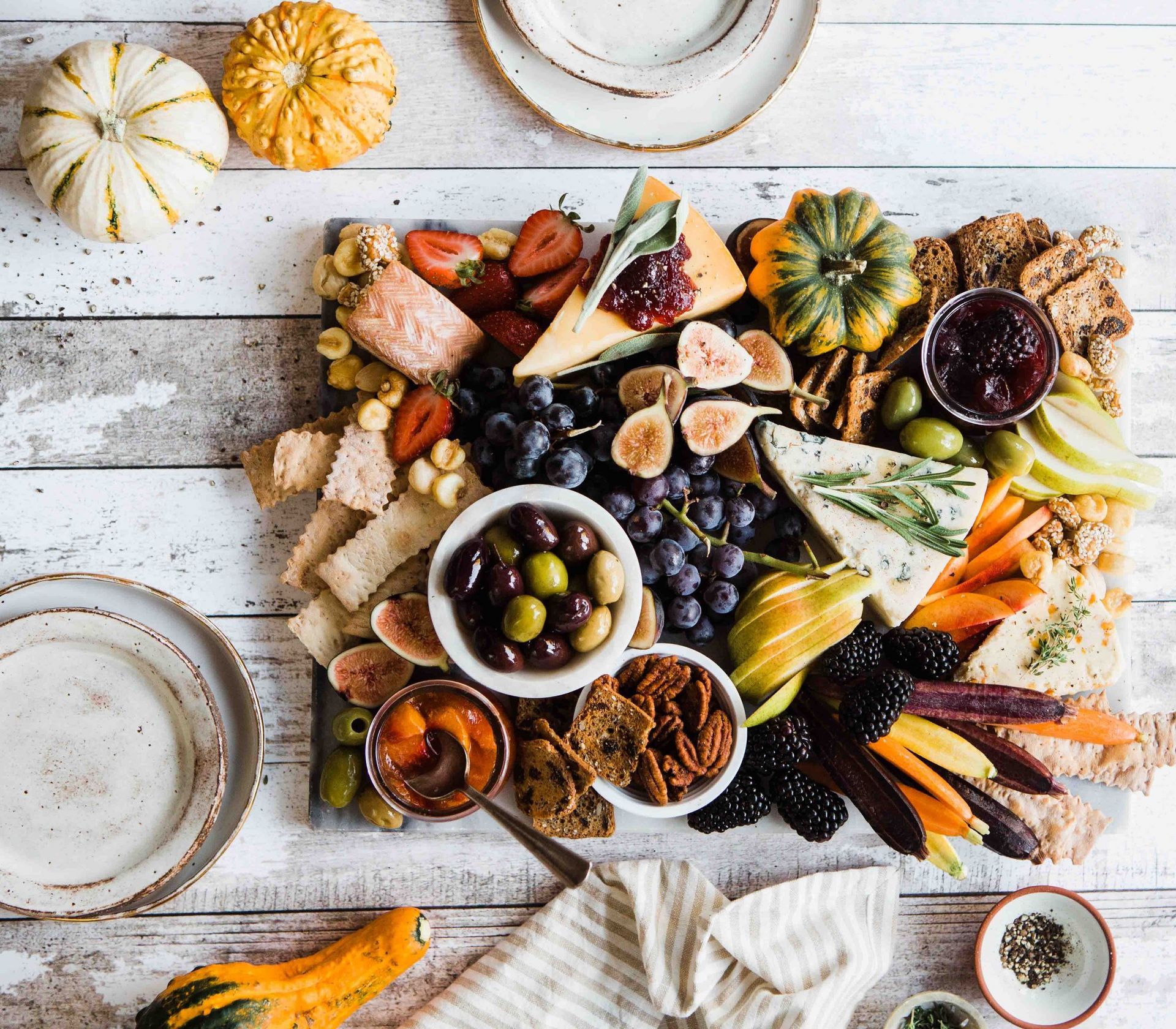 Overhead view of a colorful charcuterie board on a white wooden table with fall produce, cheese, olives, and crackers.