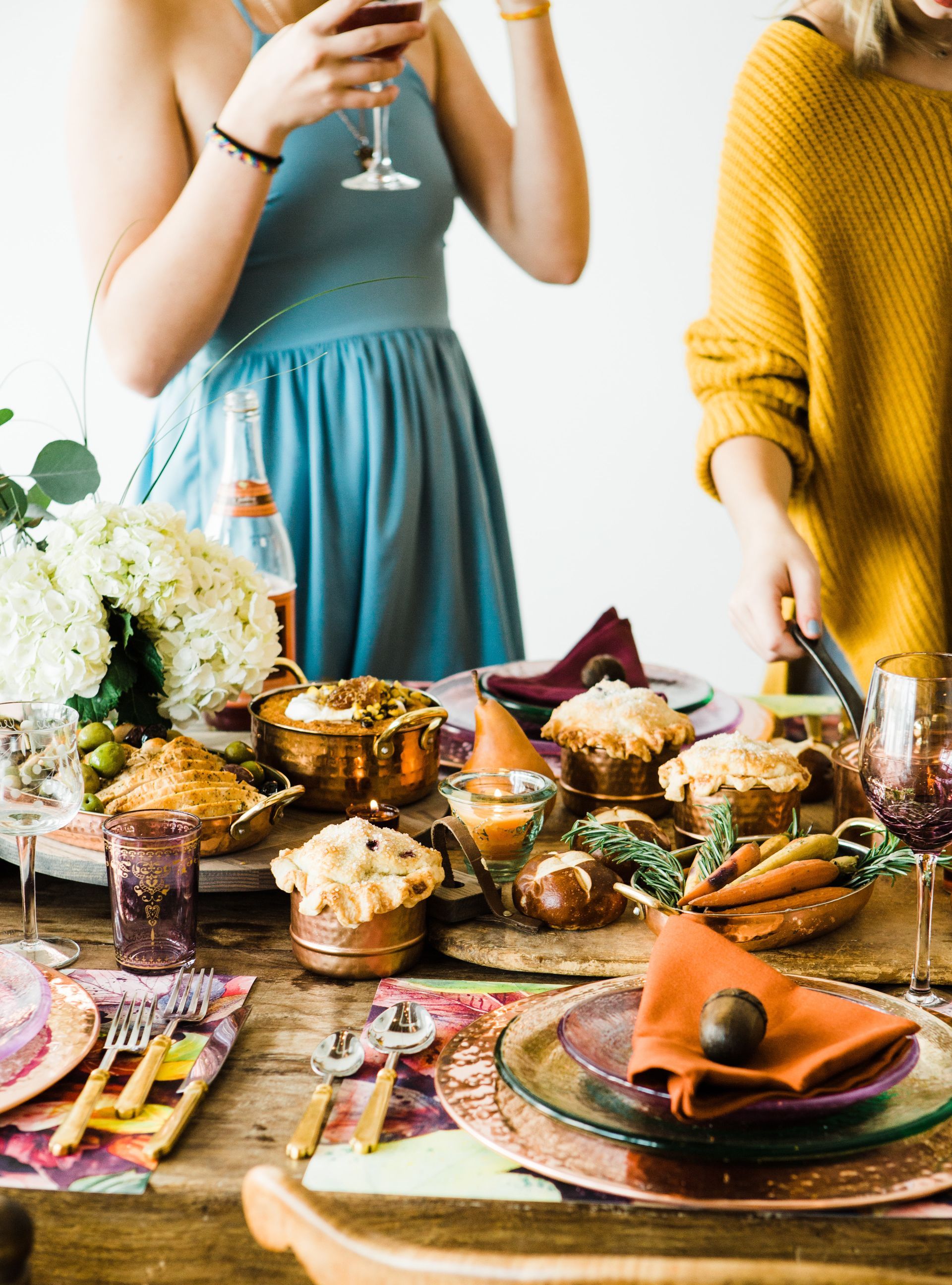 Two women at a festive table set with food, drinks, and floral arrangements. One holds a glass, the other places a dish.