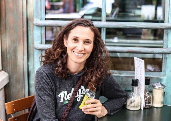 Woman with brown curly hair smiles while holding a snack bar at a cafe table outdoors. She wears a gray sweatshirt.