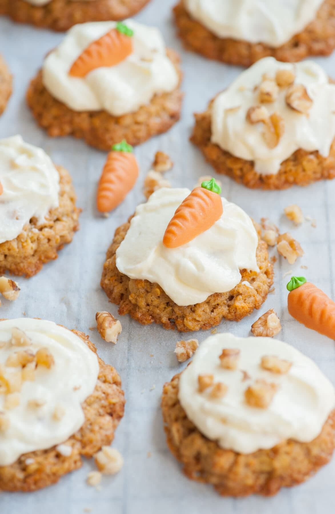 Carrot cake cookies topped with cream cheese frosting, walnuts, and a small carrot candy, on white parchment.