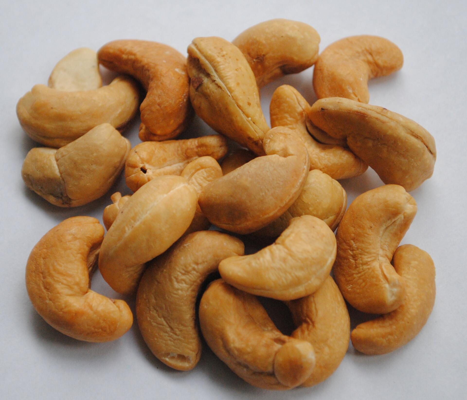 Pile of roasted cashew nuts on a white surface. Light brown and golden in color, with a curved shape.