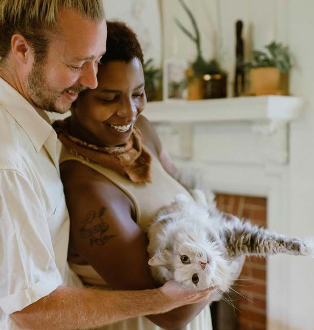 Couple smiles as they hold a fluffy gray cat indoors, in front of a fireplace.