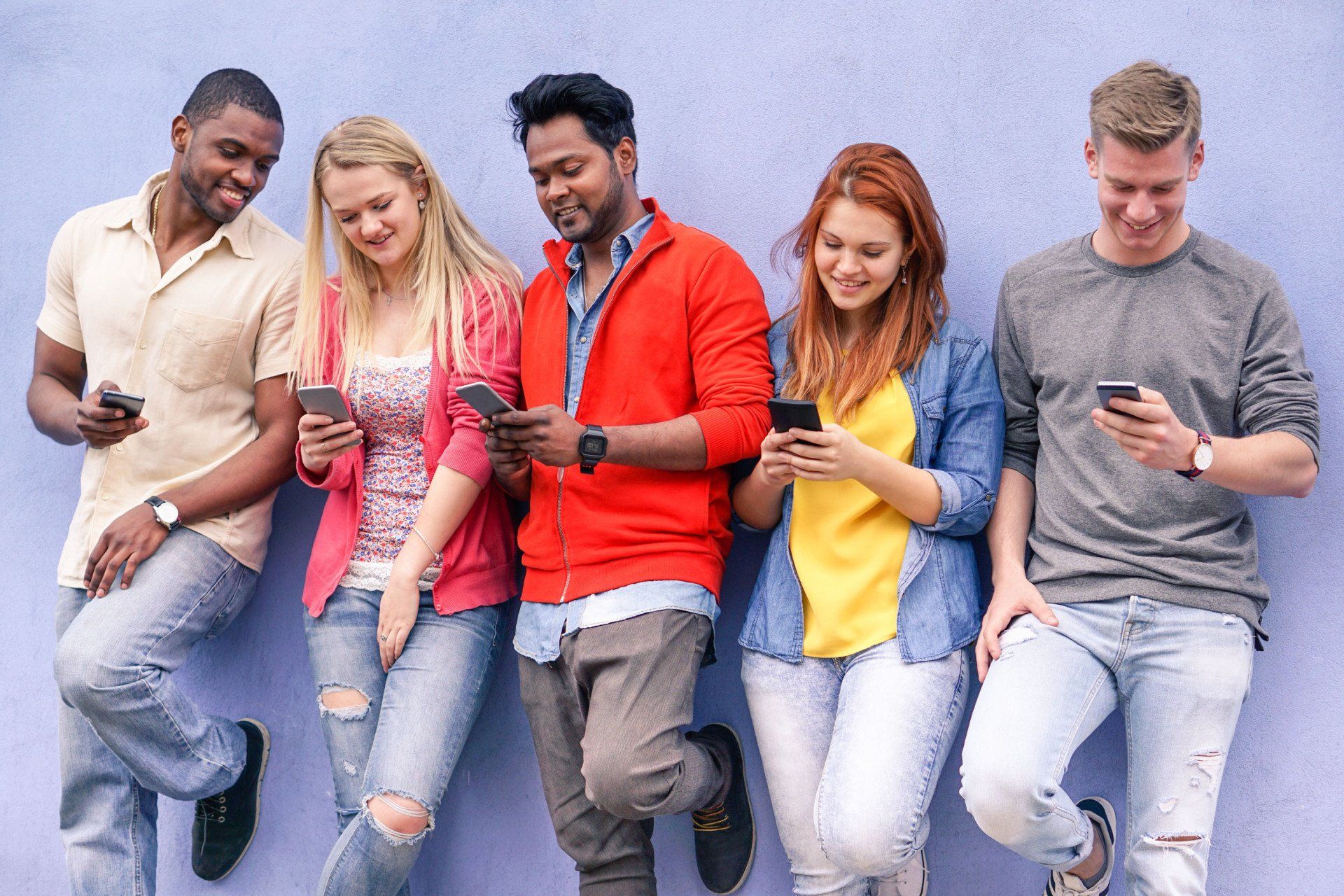Five diverse people smiling and using smartphones while leaning against a purple wall.