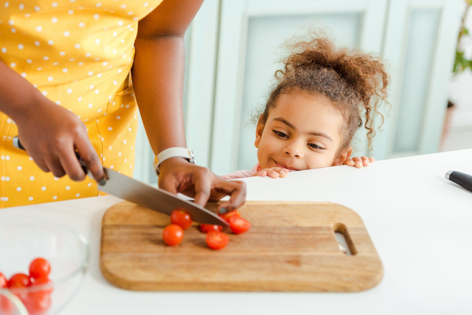 Woman in yellow apron cutting cherry tomatoes as a young girl watches, smiling, from behind a countertop.
