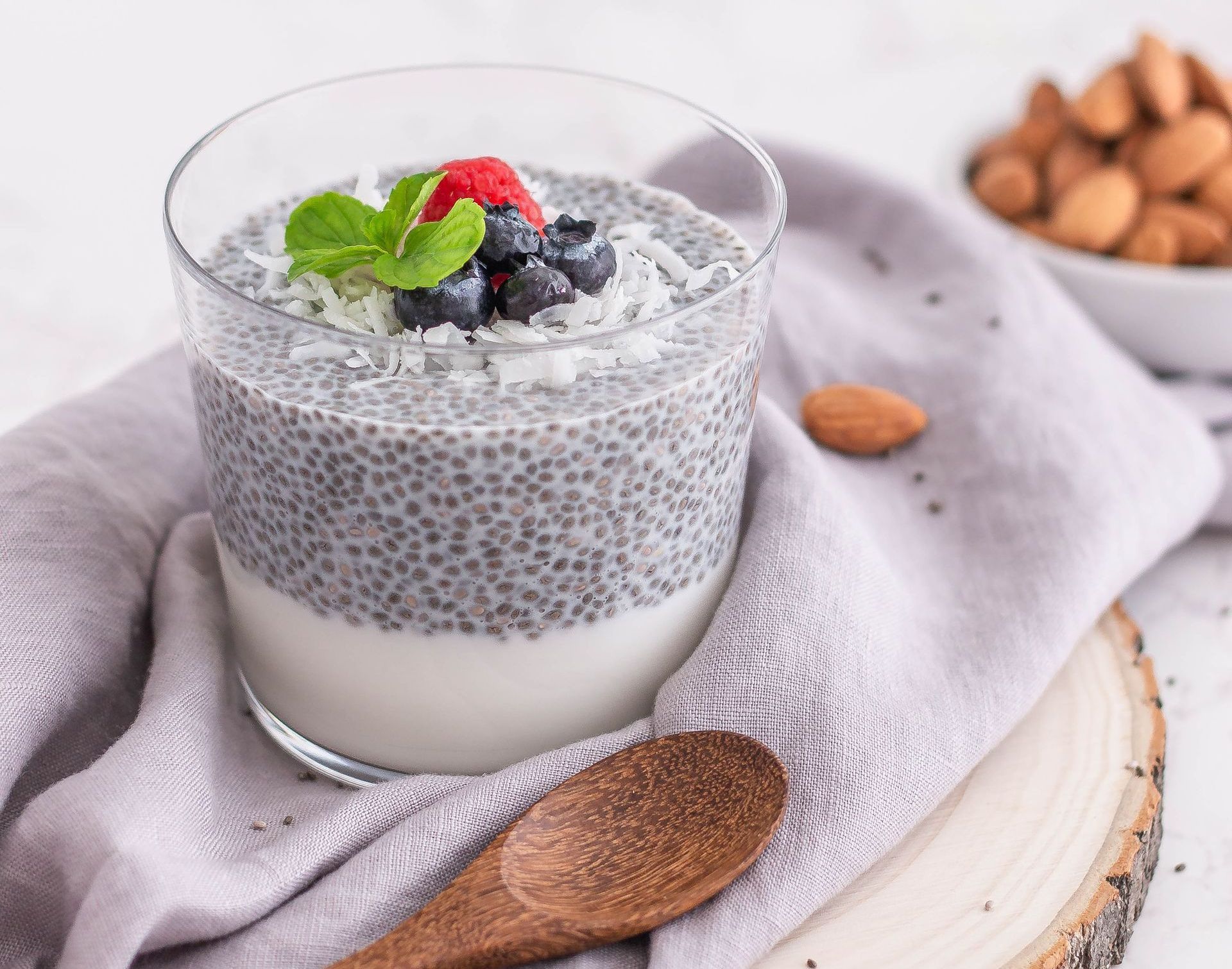 Chia seed pudding in a clear glass, topped with berries, coconut flakes, and mint. A wooden spoon and almonds are nearby.