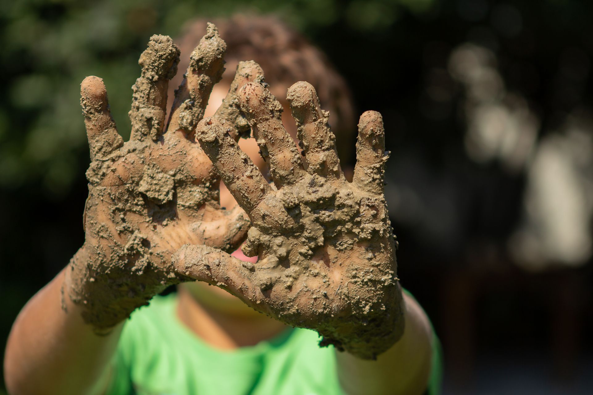Child holding up muddy hands in front of them, outdoors in green shirt.