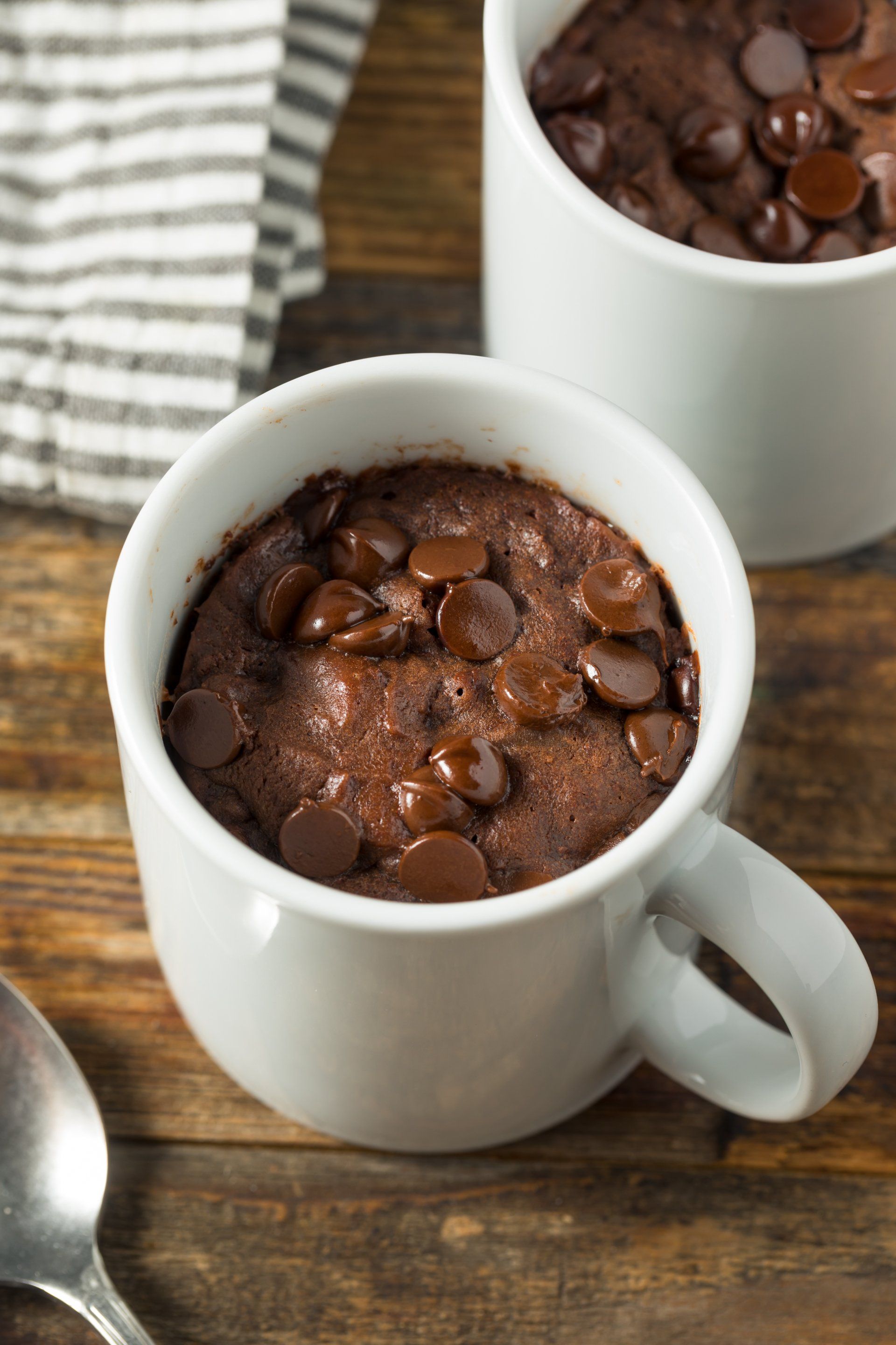 Two chocolate mug cakes topped with chocolate chips sit on a wooden table, next to a spoon.