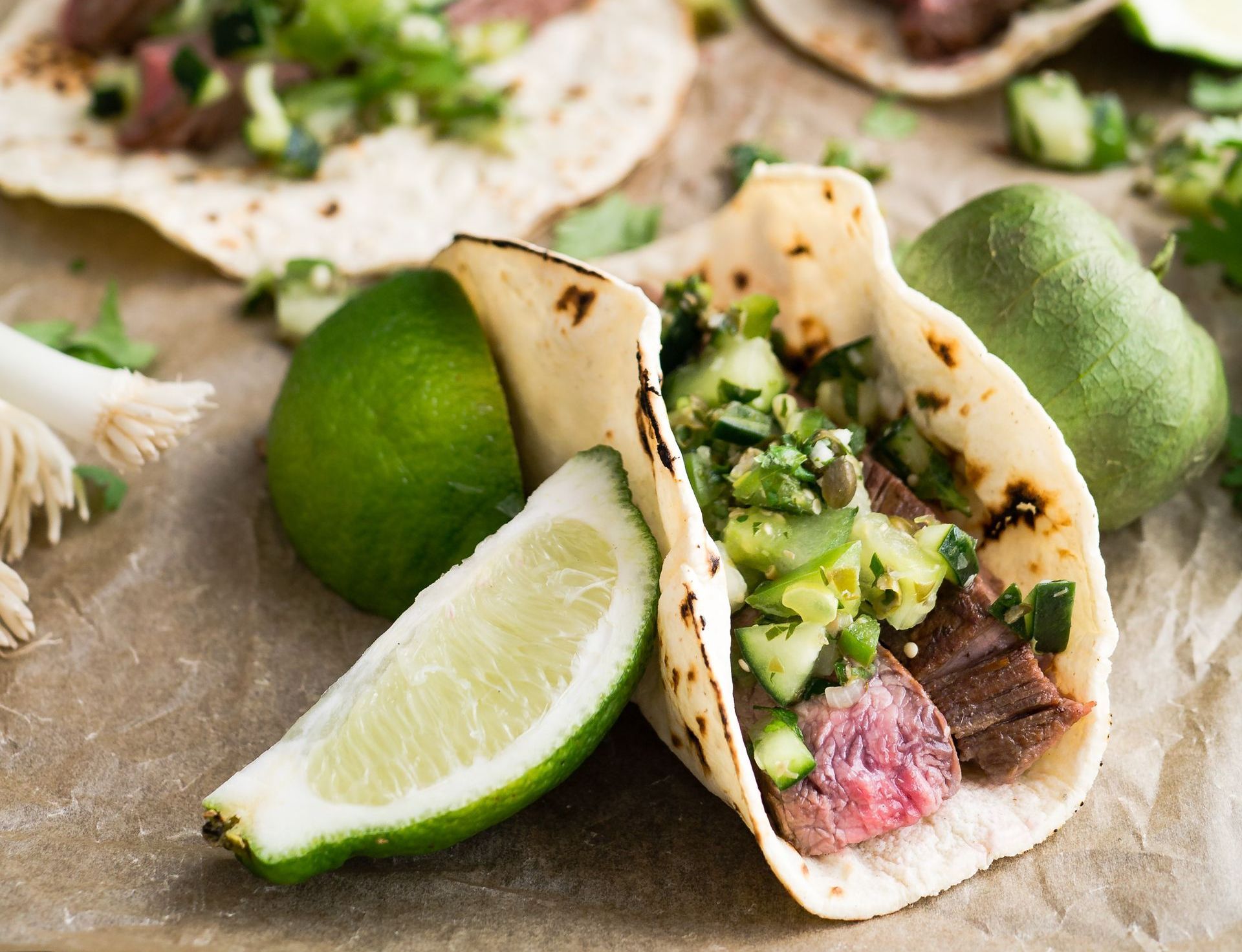 Steak tacos with fresh salsa, lime wedge, and a lime on parchment paper.