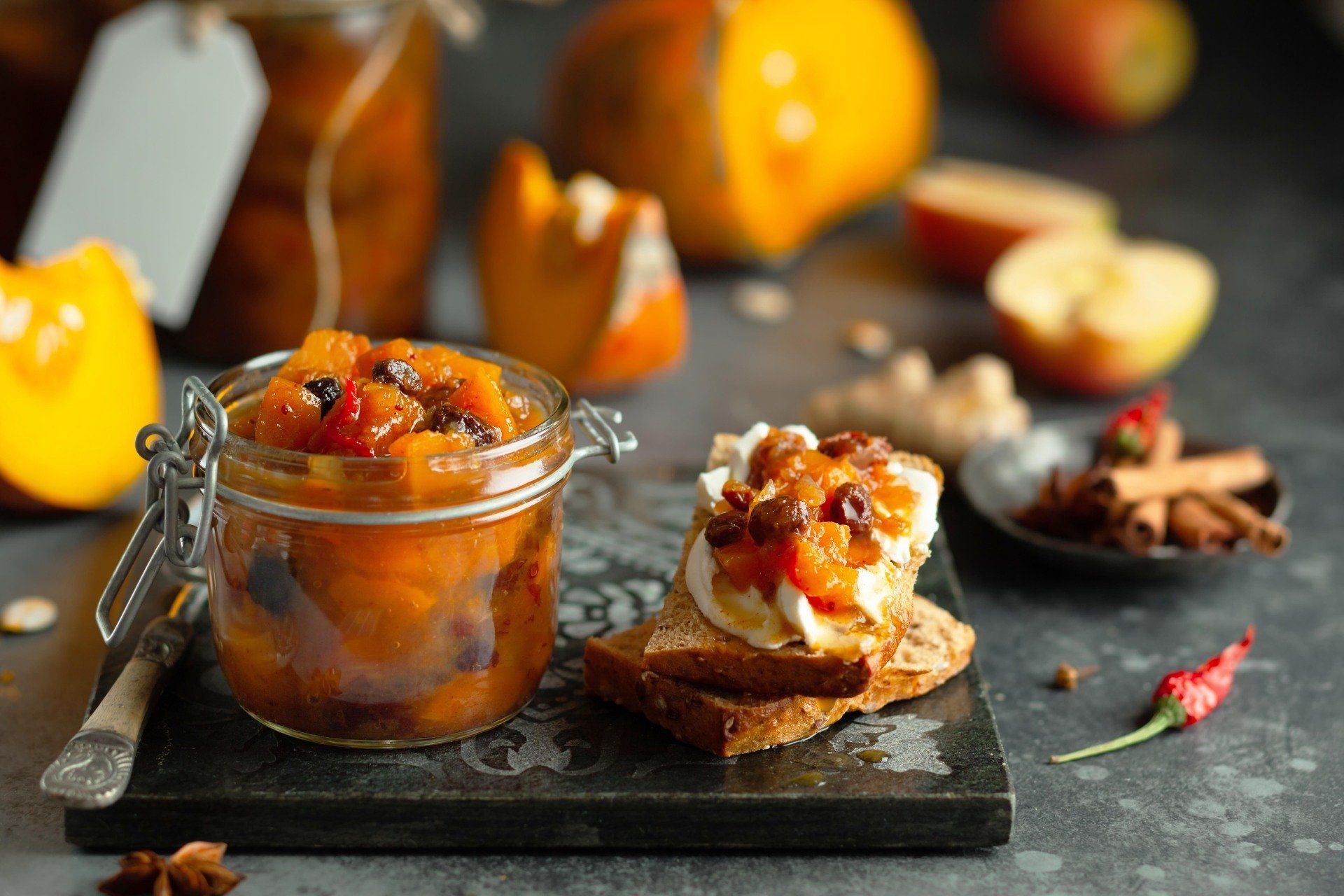 Jar of pumpkin chutney on a dark wooden board, served on toast with cream cheese, alongside spices, apples, and pumpkin.