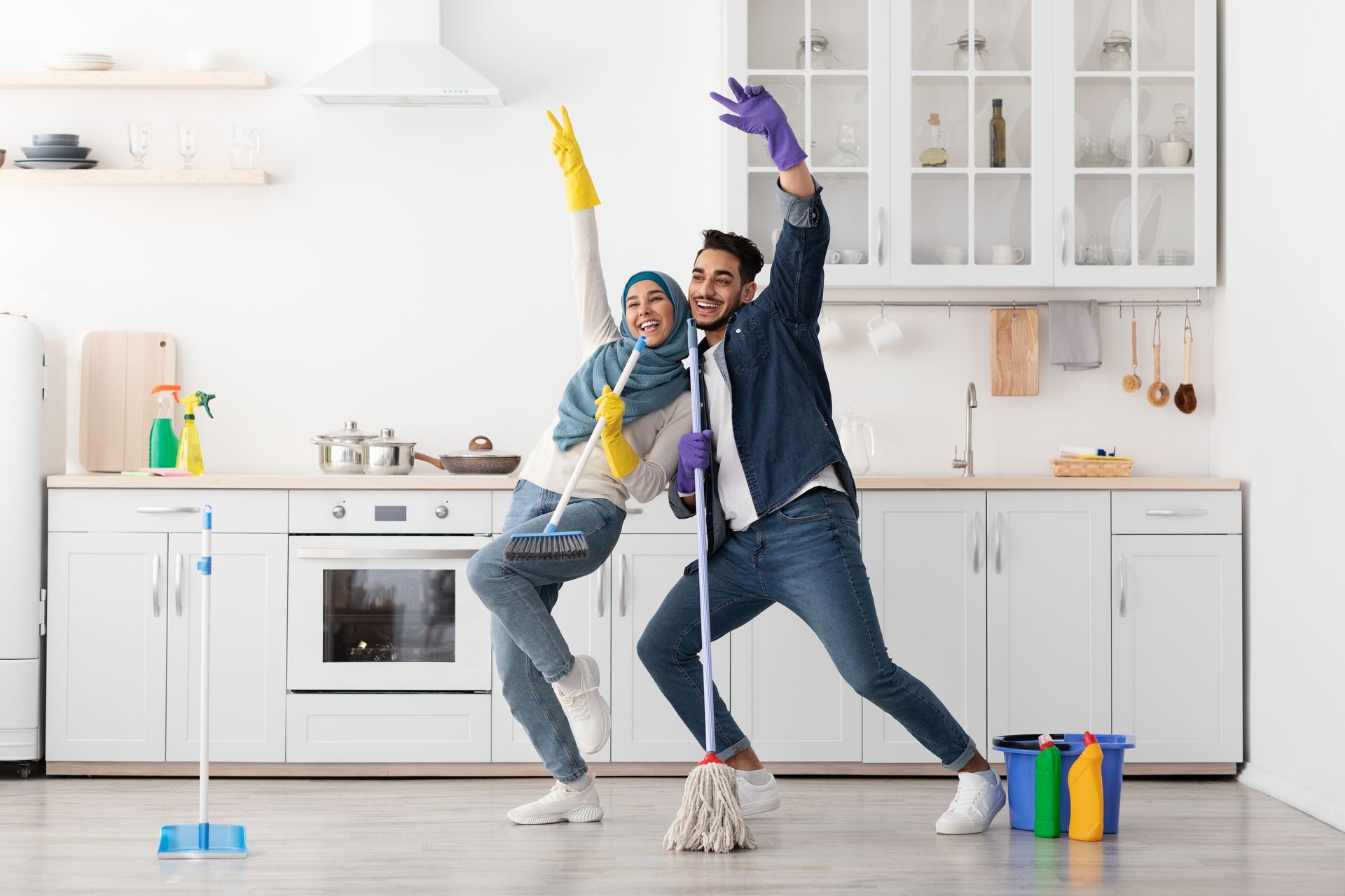 A happy couple dancing in a bright kitchen while wearing cleaning gloves and holding a mop and broom.