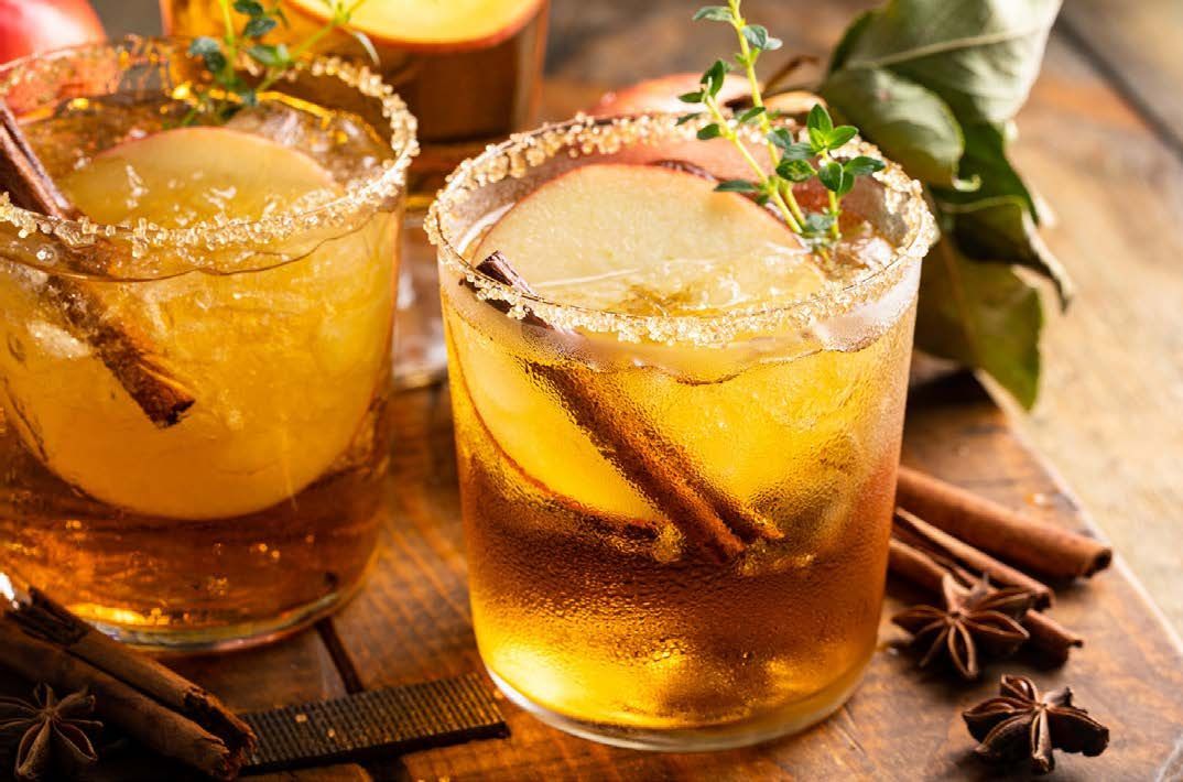 Two apple cider cocktails garnished with apple slices, cinnamon sticks, and herbs. Drinks sit on a wooden surface.