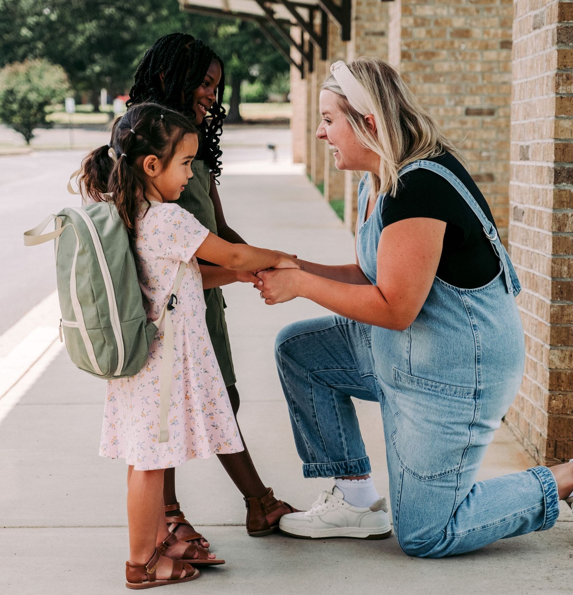 Two women and a young girl in front of a brick building. The kneeling woman holds the girl's hands.