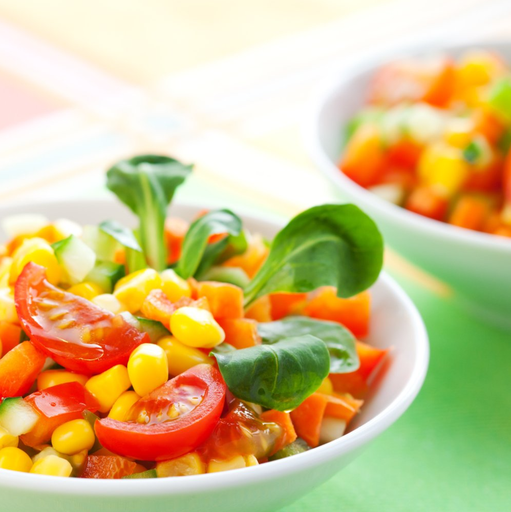 Two bowls of colorful corn and vegetable salad, including tomatoes, carrots, and greens, on a colorful table.