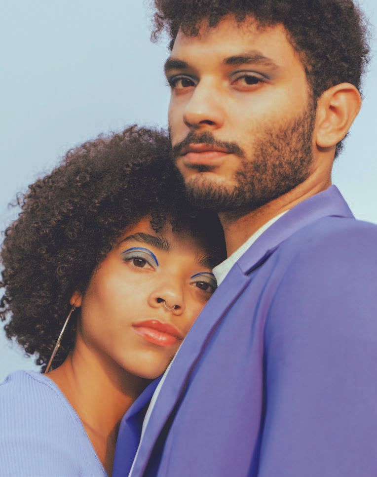 A man and a woman in matching pastel purple outfits. Woman rests her head on the man's shoulder, both with eye makeup.
