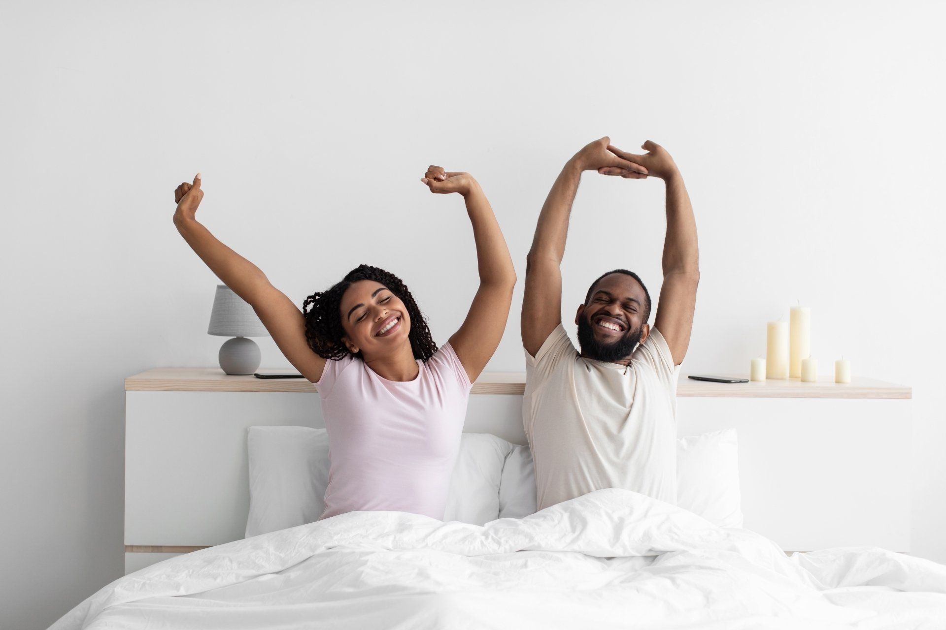 Smiling couple in bed, stretching arms up. White sheets, neutral walls, and small bedside lamp.