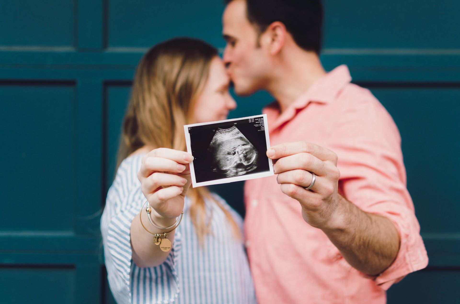 Couple holding sonogram, man kissing woman's forehead. They are standing in front of a teal door.