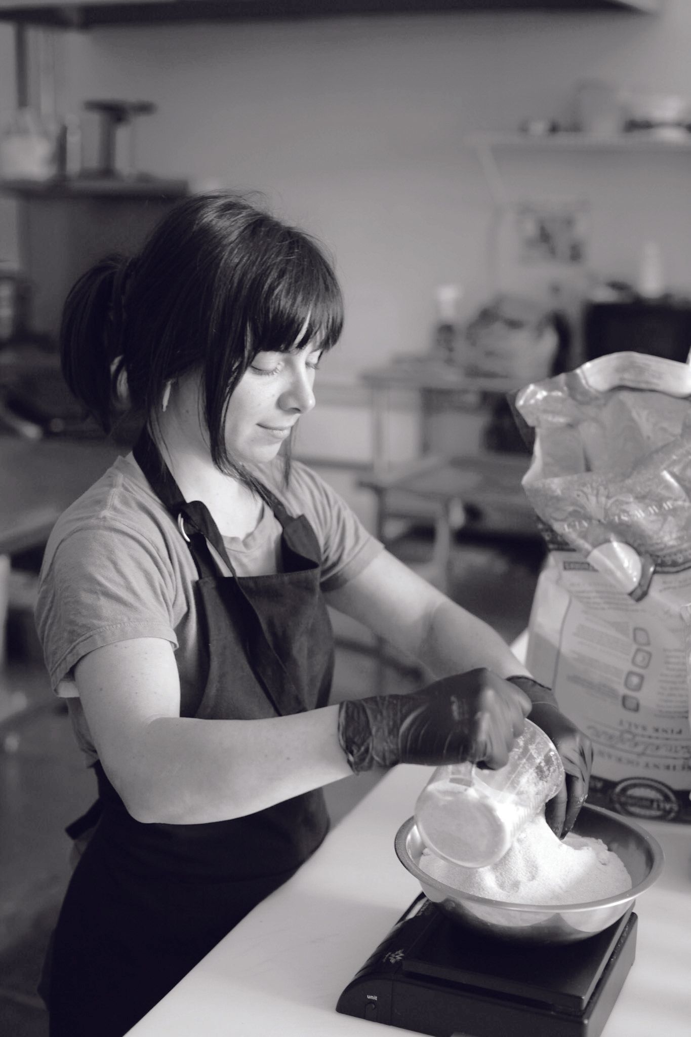Woman in apron, with short hair, measuring flour into a bowl on a scale in a kitchen.