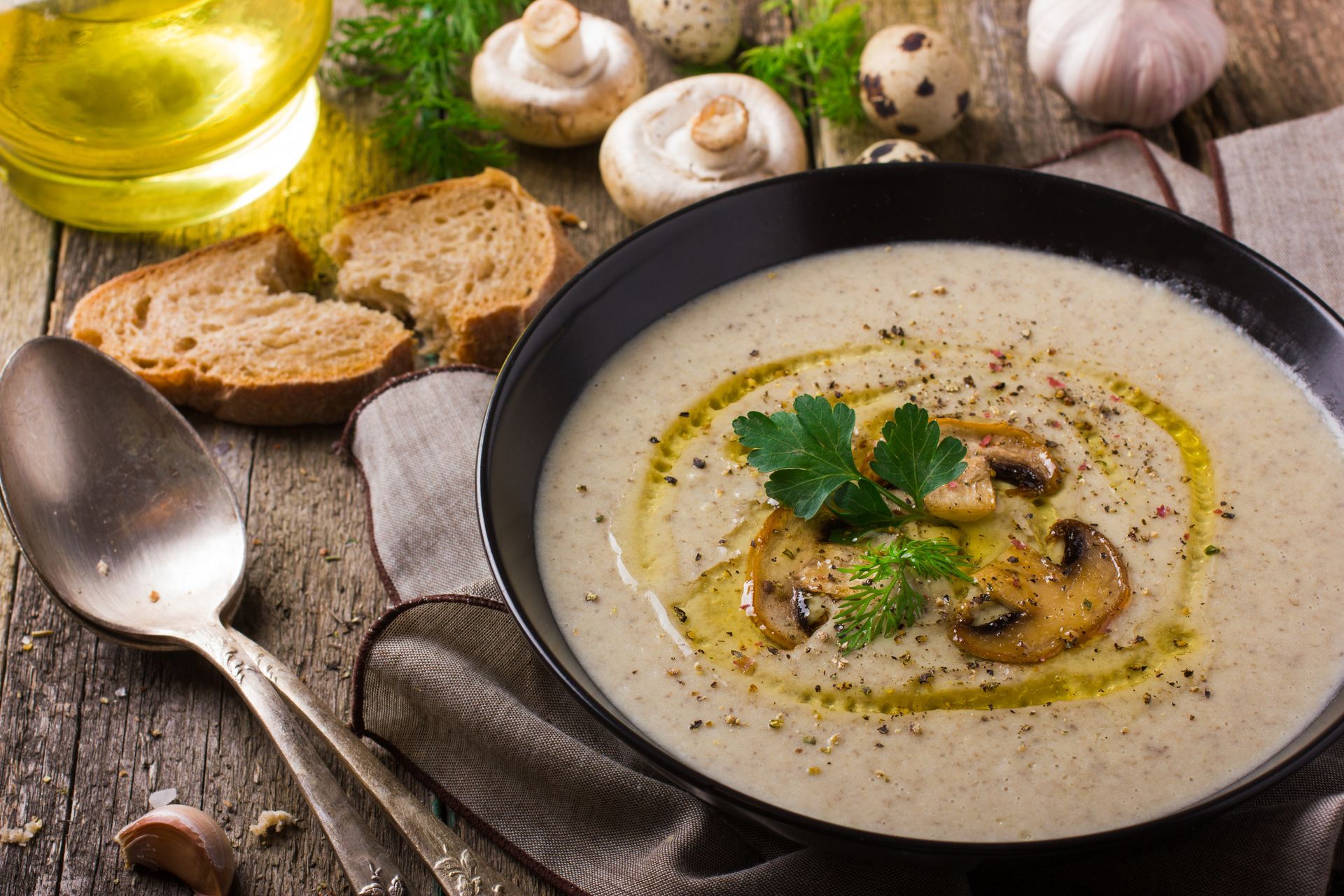 Creamy mushroom soup in a black bowl, garnished with mushrooms and parsley, with bread and ingredients on a rustic wooden table.