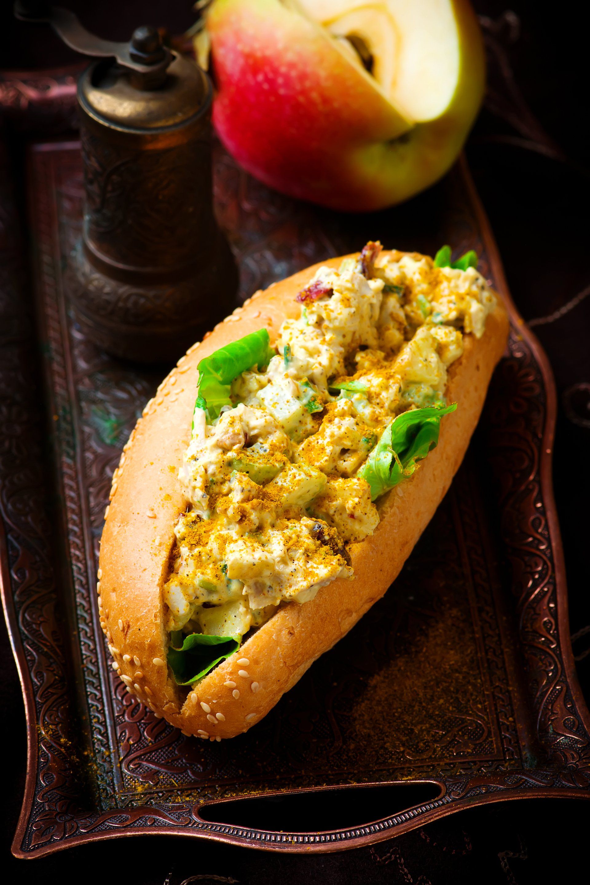Chicken salad sandwich in a sesame seed bun on a decorative tray, with an apple and pepper grinder.