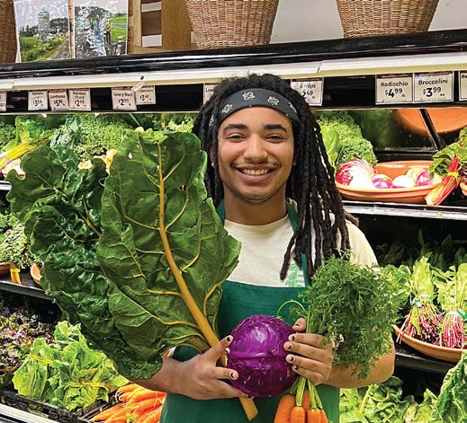 Smiling person with dreadlocks holds produce in a grocery store. They display leafy greens, a purple cabbage, and carrots.