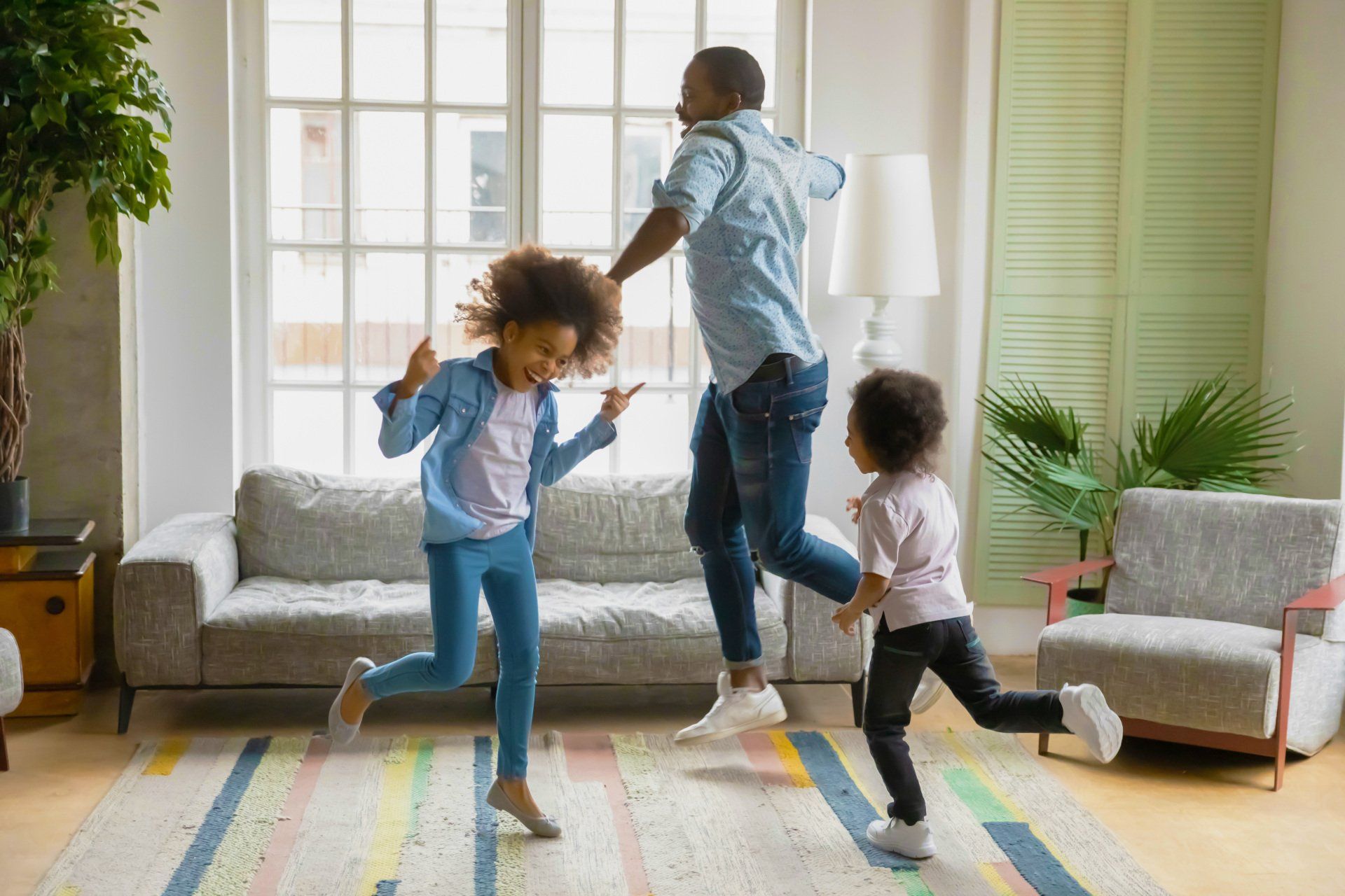 A Black father dances with his two young daughters in a living room. They are all smiling and moving excitedly on a patterned rug.