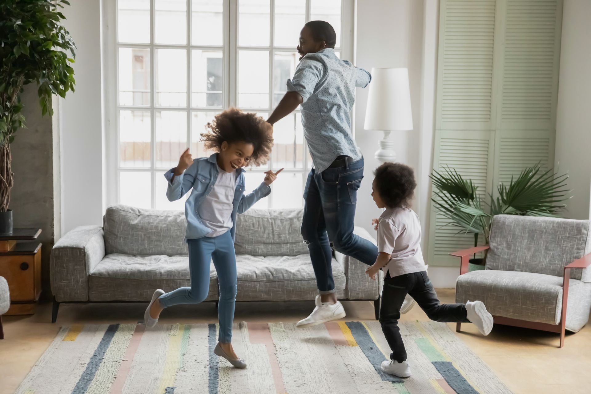 A Black father dances with his two young daughters in a living room. They are jumping with joy near a rug.