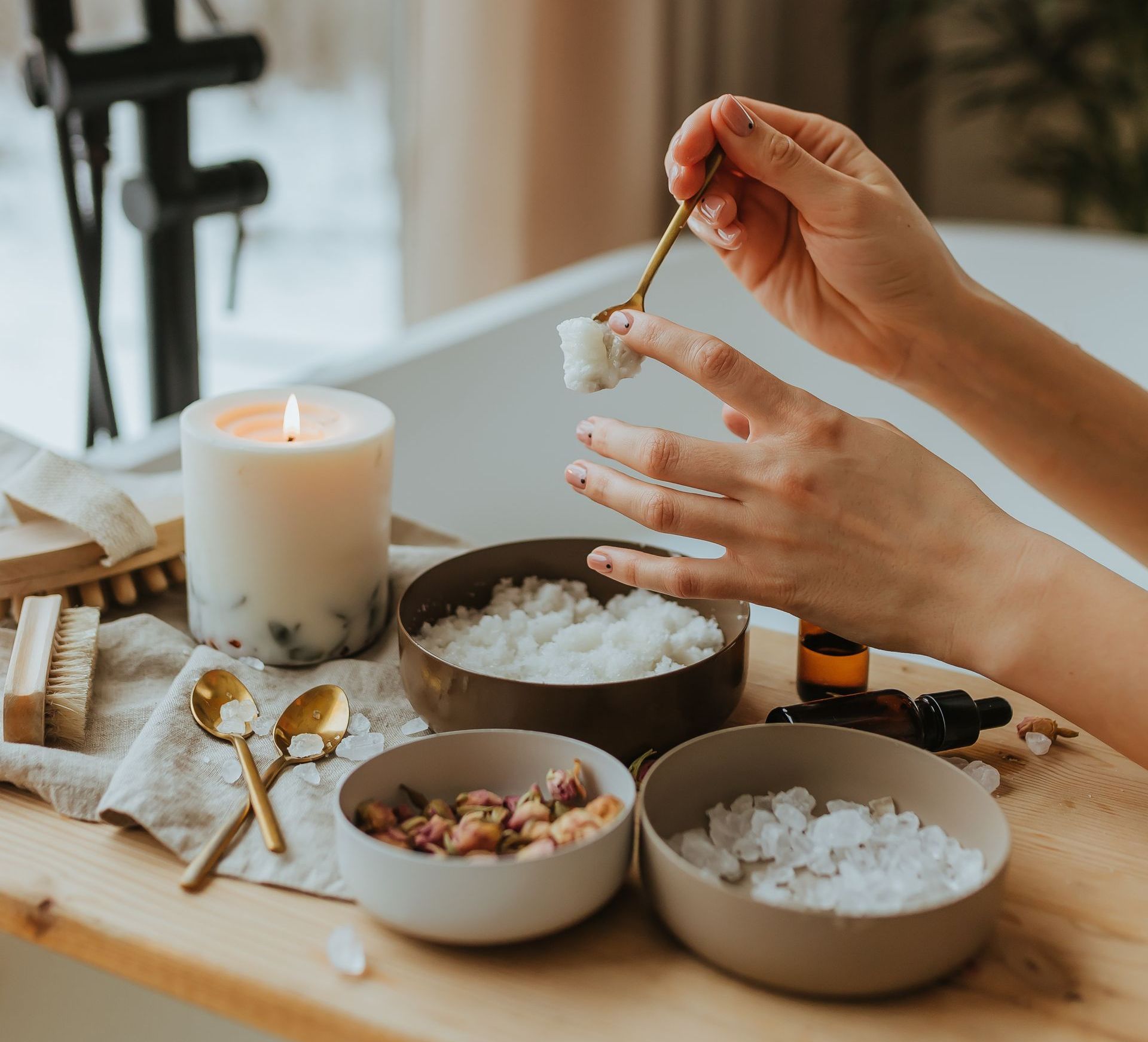 Person preparing a bath with bath salts, candle, and essential oils. The setting is a spa-like bathroom.