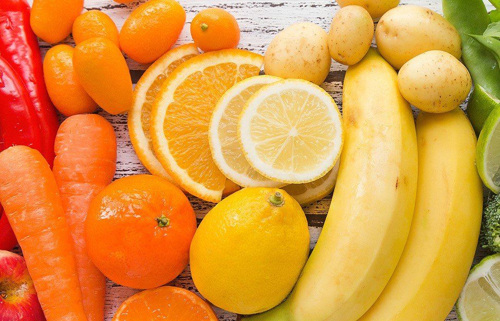 A vibrant rainbow of fruits and vegetables: red pepper, carrots, orange, lemon, banana, and more, arranged on a light wood surface.