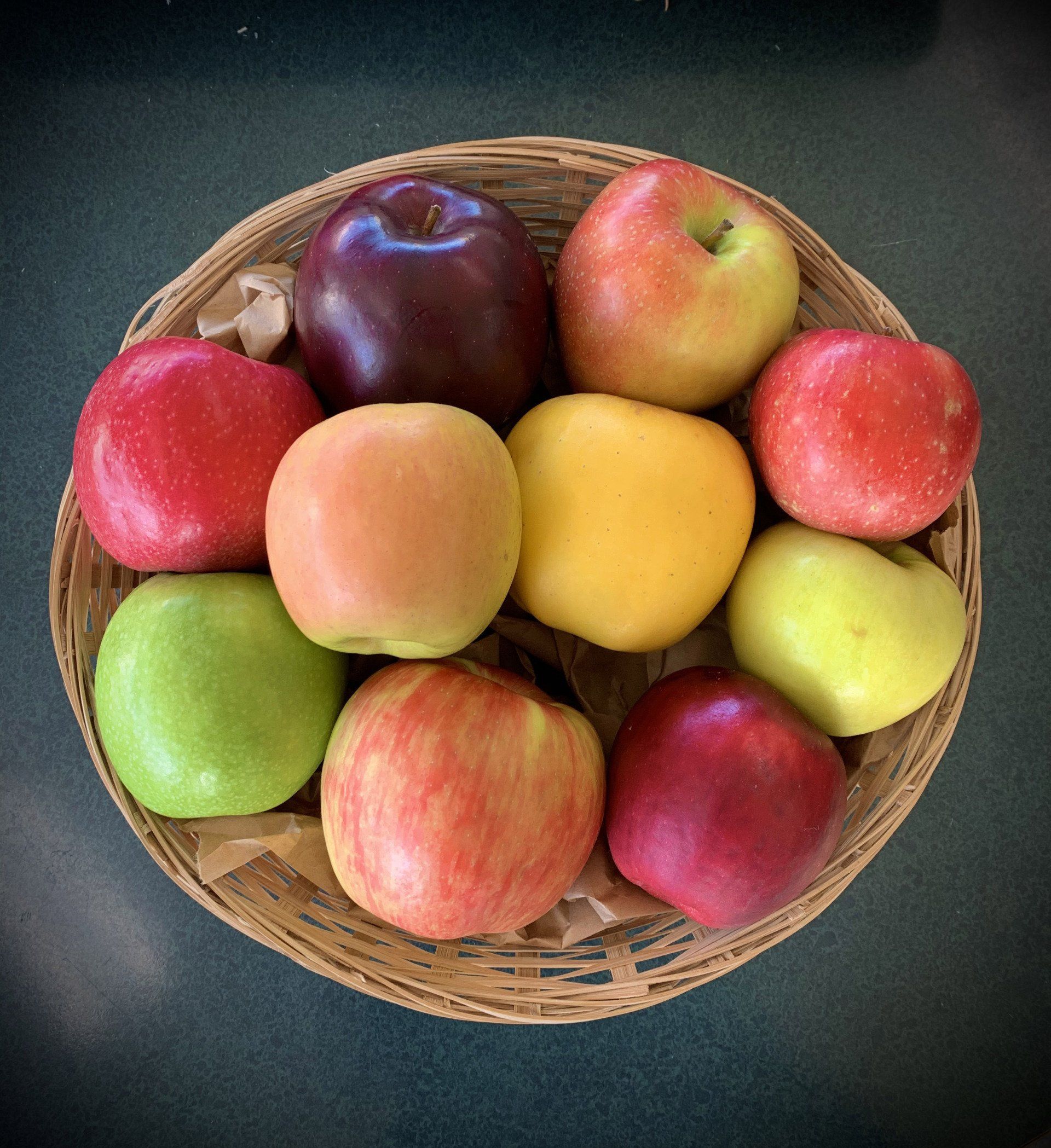 A wicker basket filled with assorted colorful apples: red, green, yellow, and purple.
