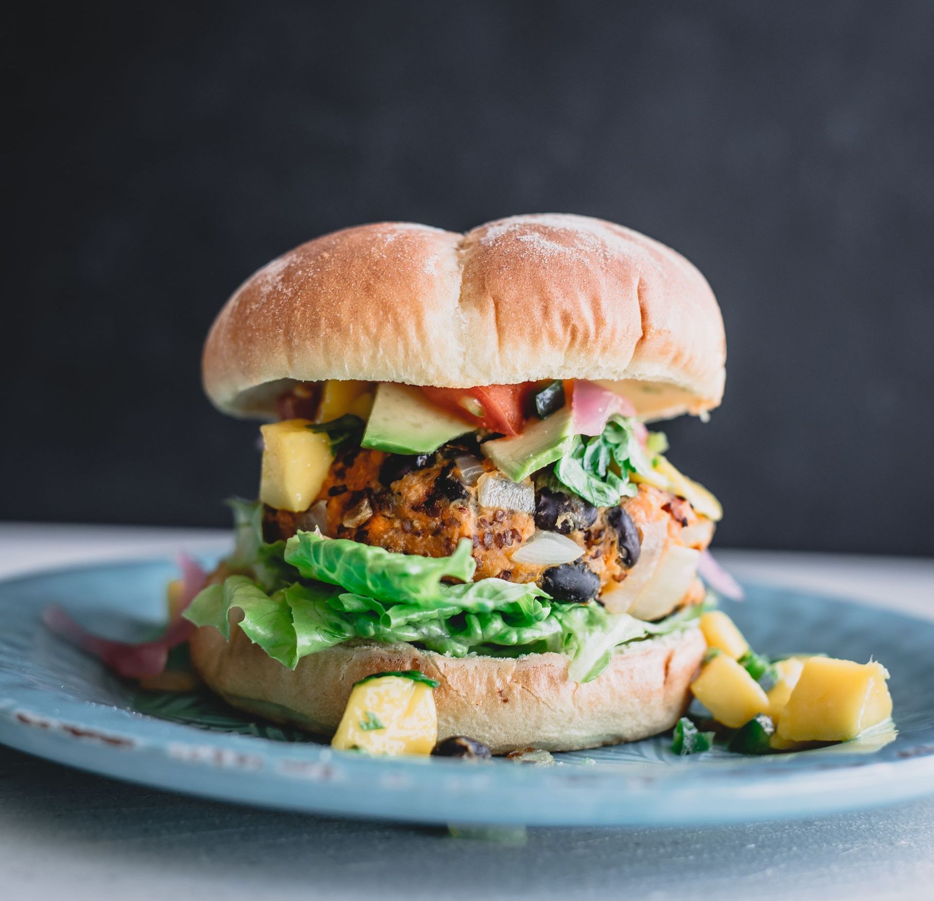 Vegan burger with a black bean patty, topped with mango, lettuce, and red onion on a bun, on a blue plate.