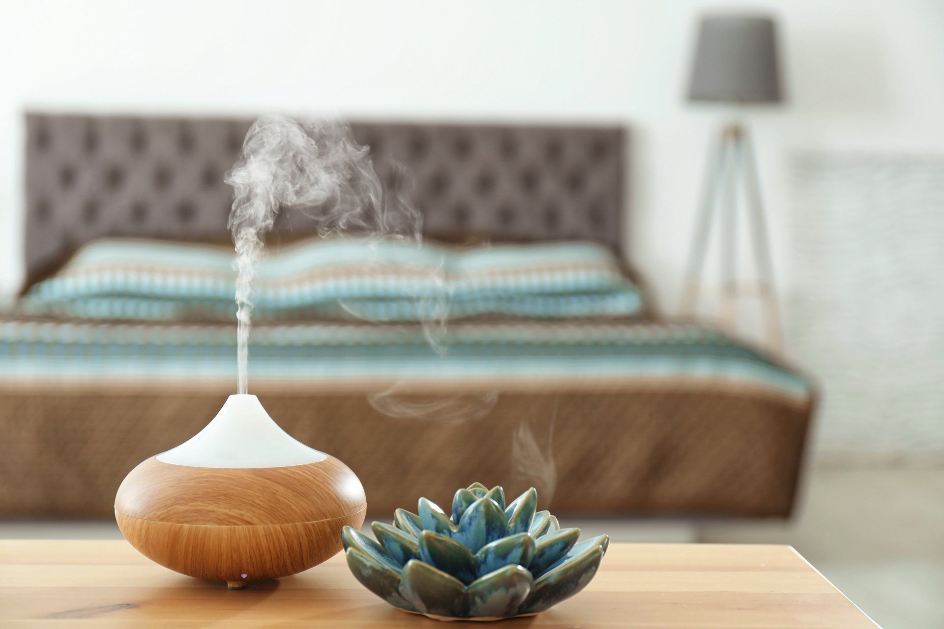 An essential oil diffuser releases steam on a wooden table in a bedroom with a bed and floor lamp in the background.